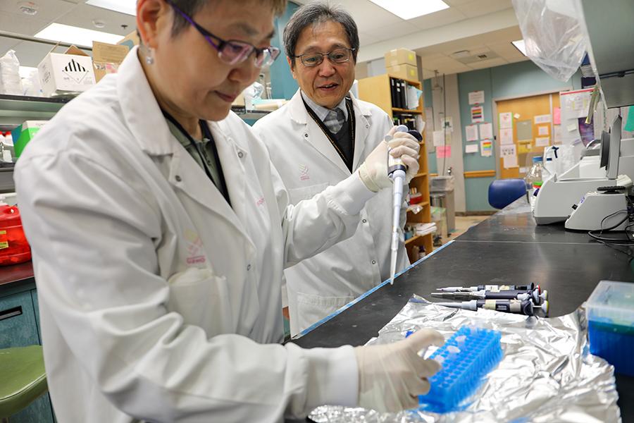 A researcher in a lab holds a liquid dropper over test tubes as another researcher looks on.